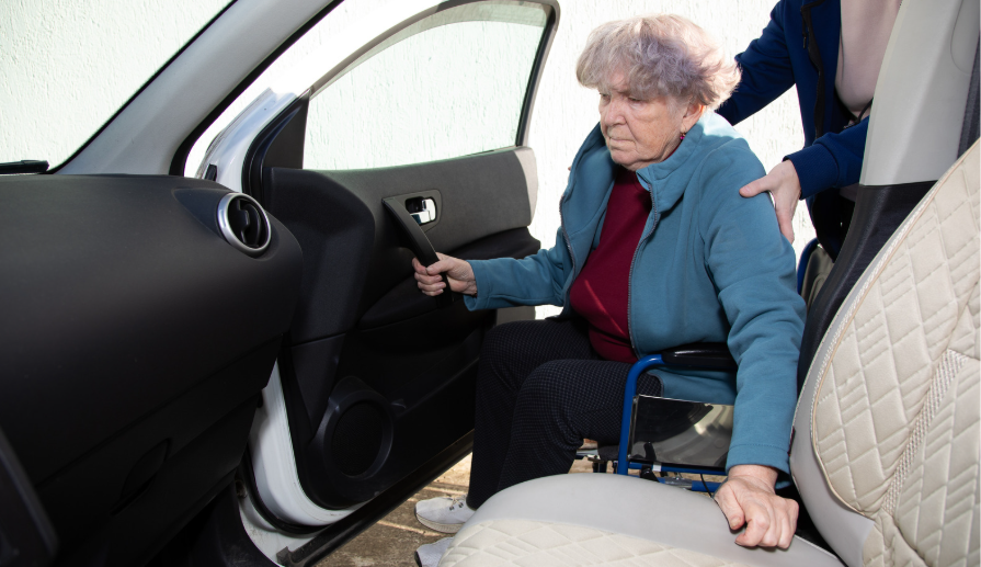 Older adult transferring from a wheelchair into a vehicle with assistance from a caregiver.